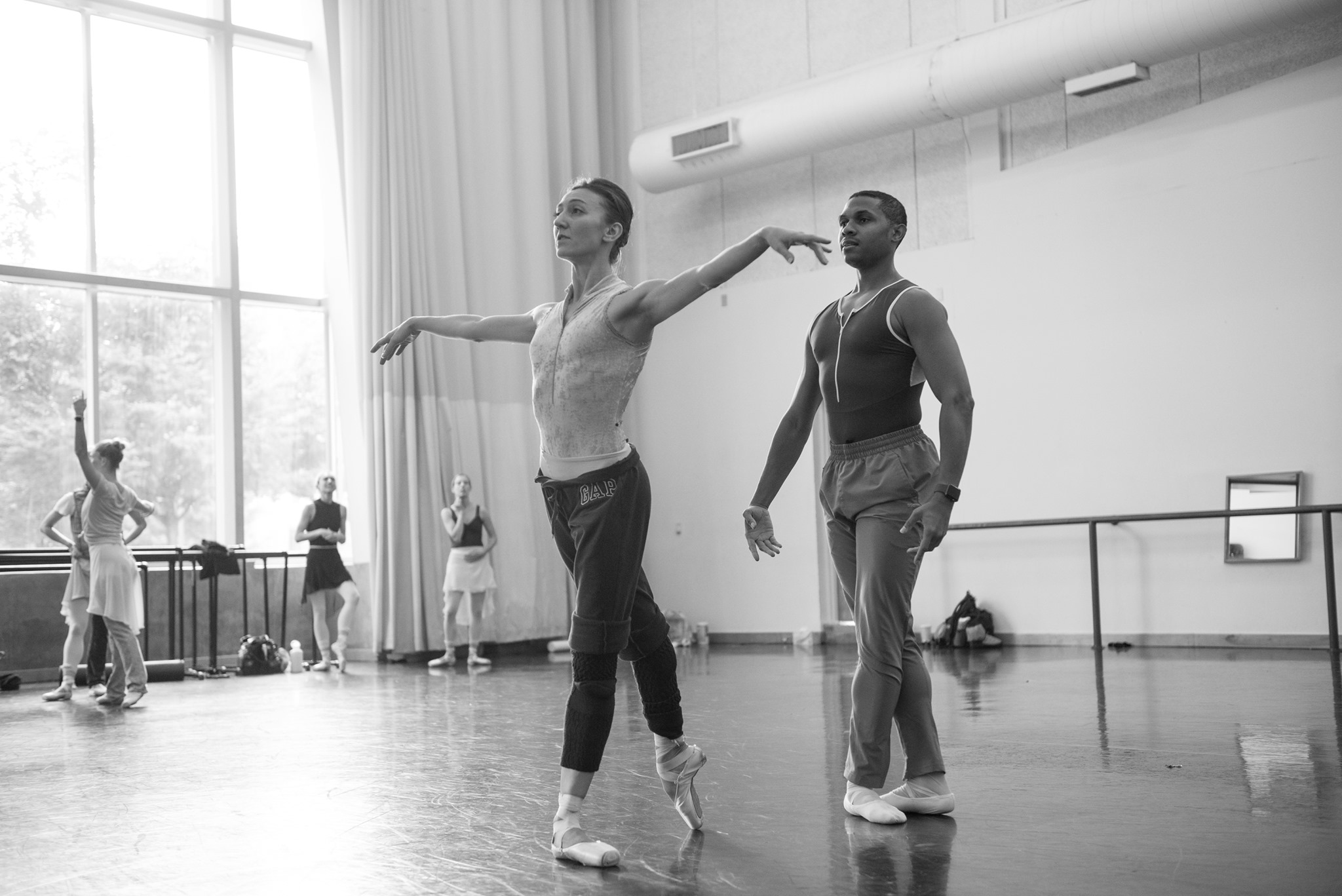 A black and white photo featuring two ballet dancers rehearsing in a studio. Other dancers work behind them.