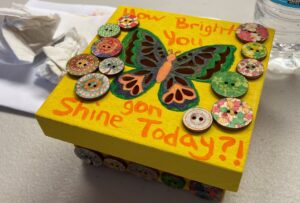 A small yellow box covered in buttons, with a butterfly painted on it and the phrase "How Bright You gon Shine Today?!"