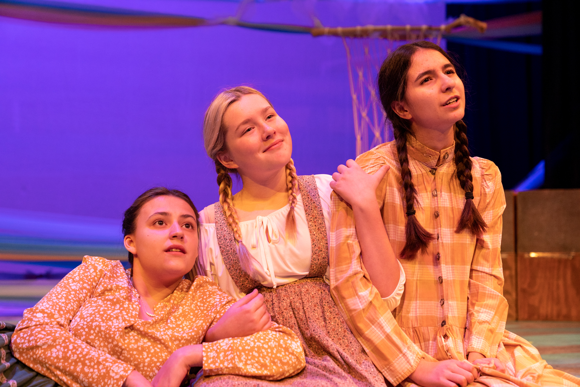 Three young high school girls sit, lit with theatrical lighting, arms linked and looking out wistfully as part of a scene in "The Selkie Wife."