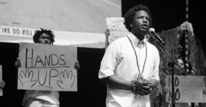 A black and white photo of Brandon Harrison speaking at a microphone, behind him, a man holds a handwritten sign saying "Hands Up."