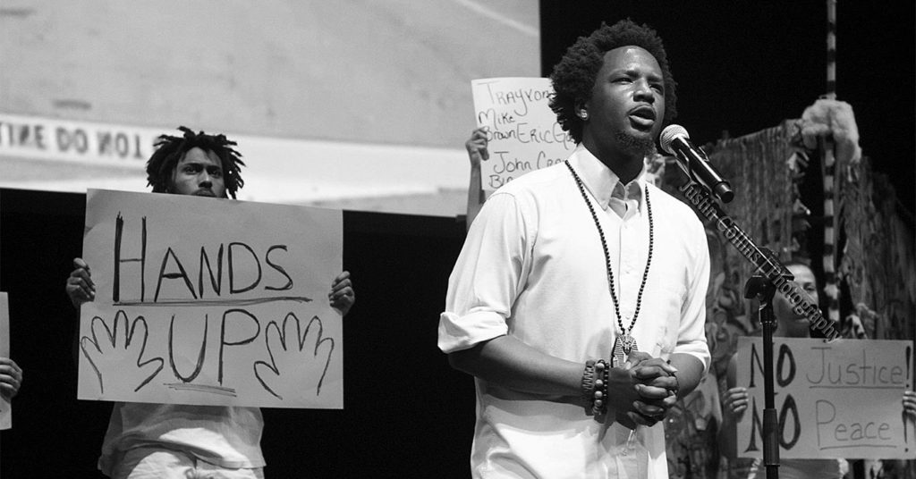 A black and white photo of Brandon Harrison speaking at a microphone, behind him, a man holds a handwritten sign saying "Hands Up."