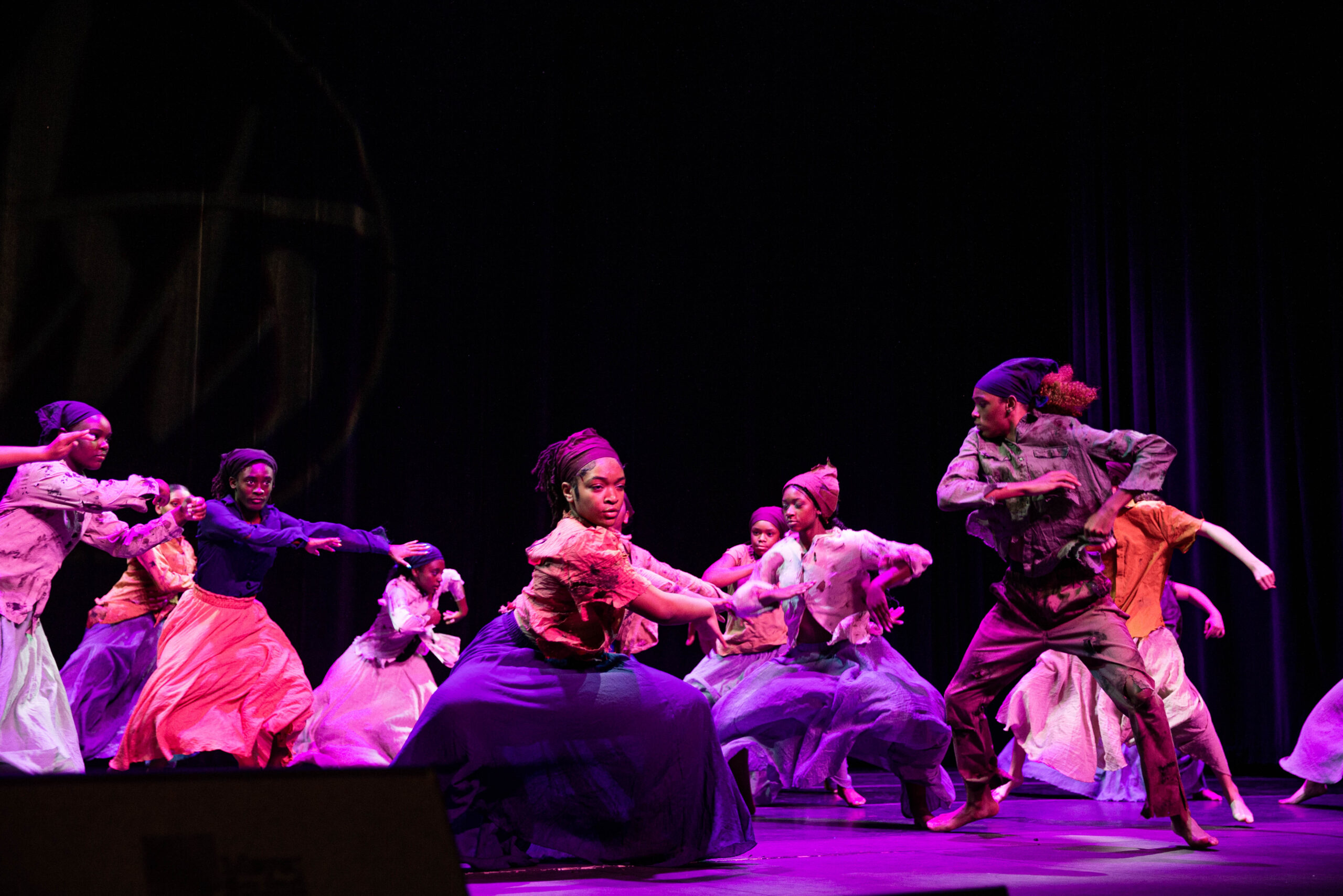 A group of performers in the midst of a dance, lit with purple light. They are performing a piece called "Harriet," inspired by Harriet Tubman.