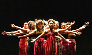 A group of girls stand onstage in matching red dresses, theatrically lit in a calm and powerful choreographed formation, arms outstretched.