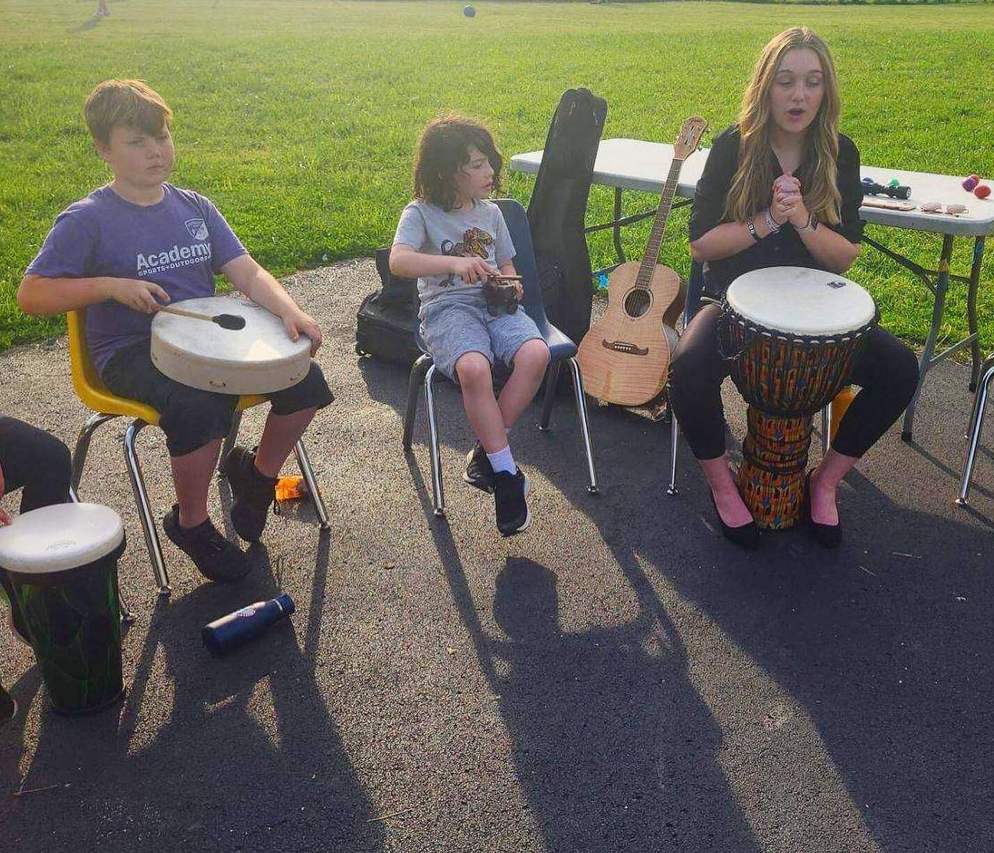 A woman sits outside at a chair with a large drum. Two kids are beside her holding other drums. A guitar leans against a table behind them.