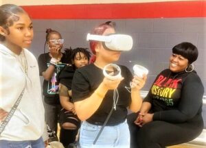 A woman smiles, looking on at a girl holding a VR headset, while three other kids stand behind her.