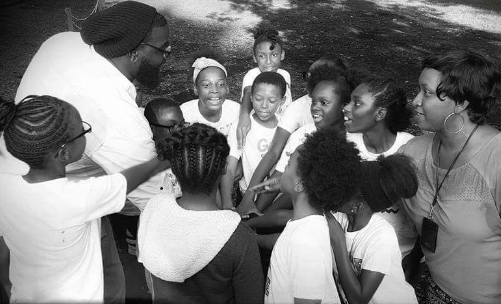 A Black and white photo of two teachers and about 10 smiling kids and youth in a close circle, hands placed into the center as though about to cheer.