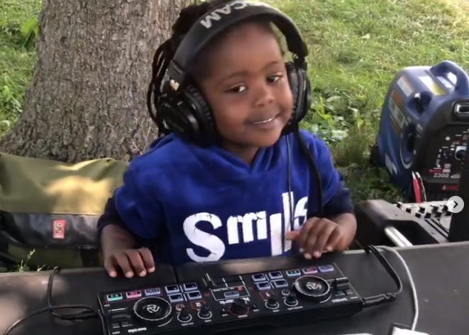 A young kid sits at a table outside, smiling happily, wearing a large set of headphone, with a piece of DJ equipment the size of a keyboard in front of them.