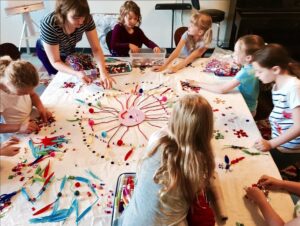 A teacher with a group of young students around a table, using objects and art supplies to make colorful circular patterns featuring a smiling face at the center.