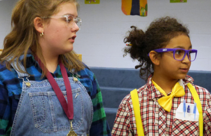 Two gradeschool students performing as part of a play in a school library, dressed as nerds with glasses and suspenders.