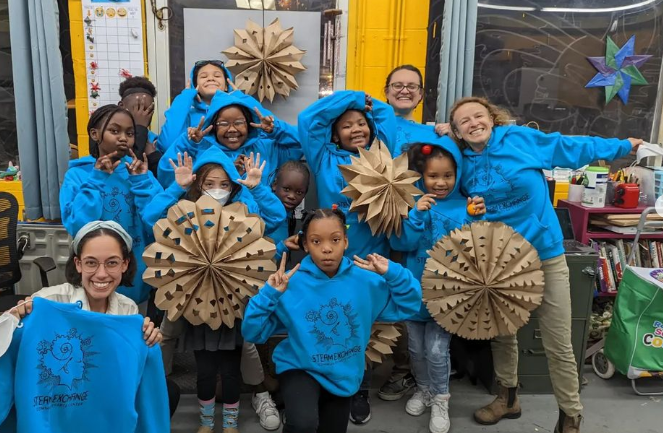 A couple teachers and a group of smiling kids in matching bright blue hoodies pose for a picture with four large handmade brown paper snowflakes.