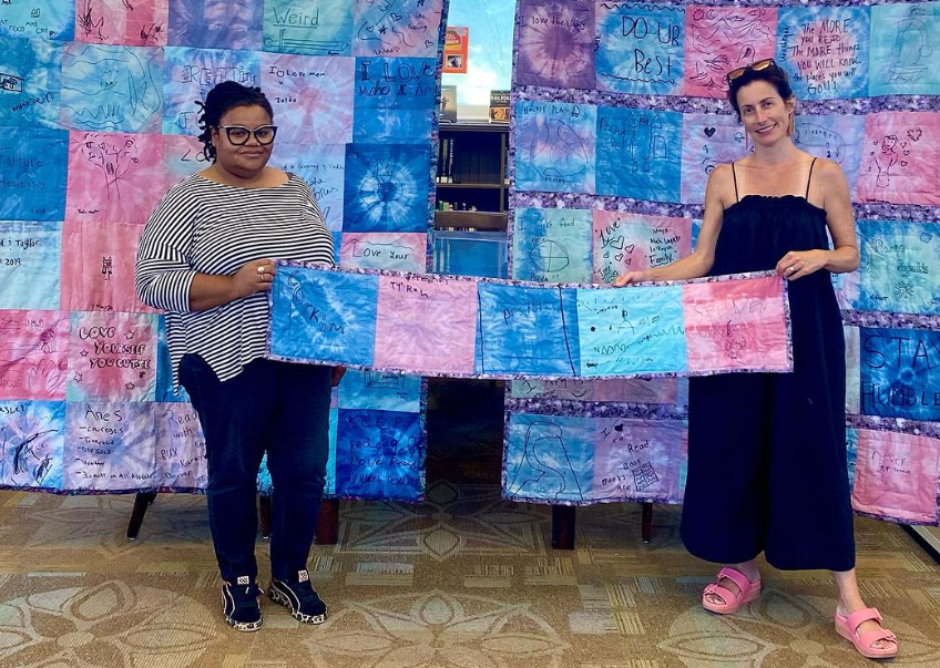 Two women hold a row of five tie-dyed quilt panel. Two quilts in matching pinks, blues, and purples are stretched behind them.
