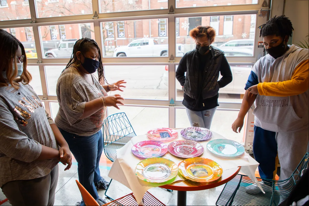 Four people stand around a table with colorful plates set out, one actively explaining something to the others as they all look at the plates.