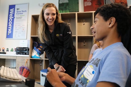 A smiling woman talks to a teen boy sitting in the teen tech center.