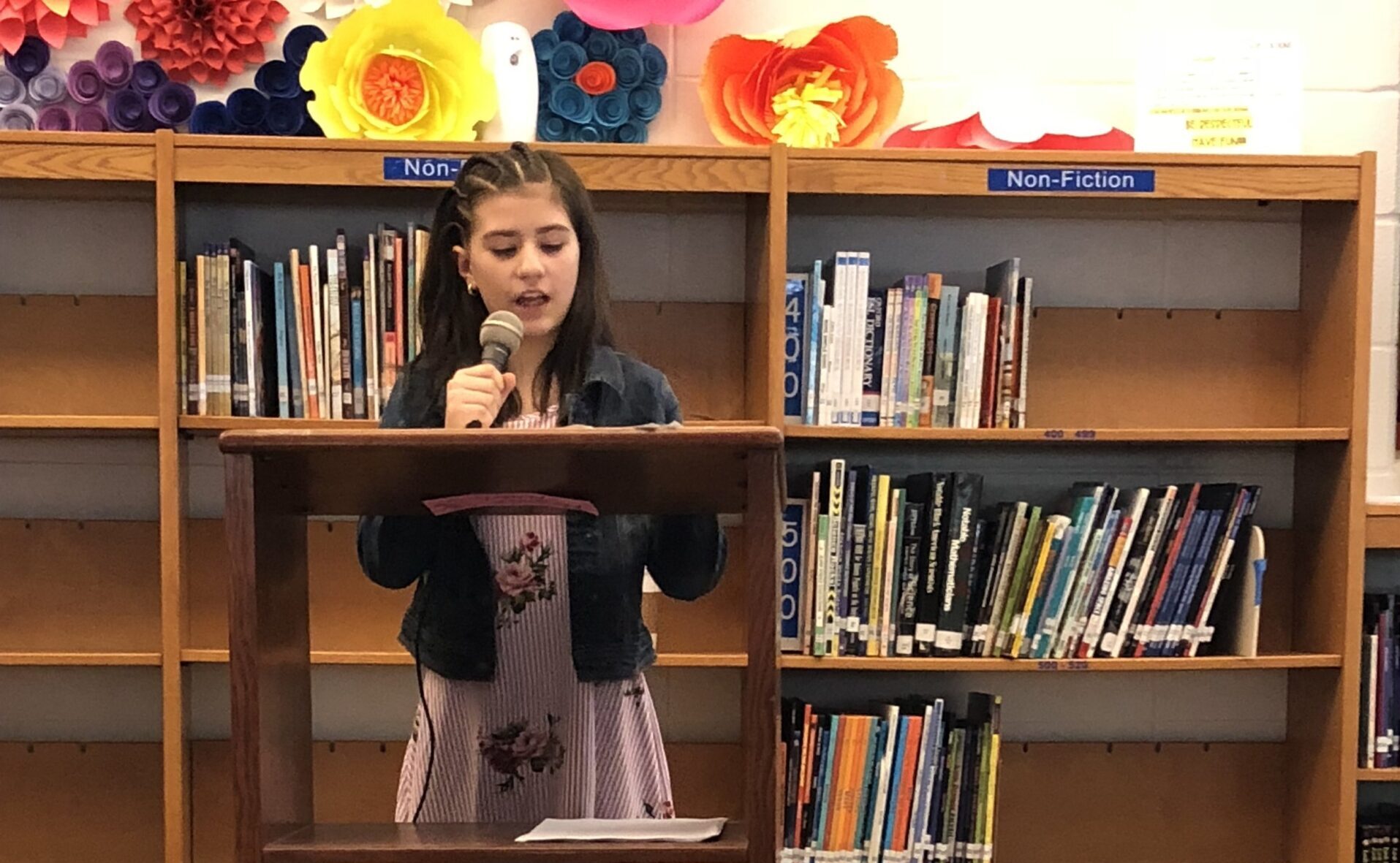 A kid stands at a wooden podium in front of school bookshelves, holding a microphone and reading from a paper on the podium.