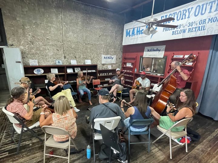 About a dozen adults sit on fold-out chairs in a close circle, playing musical instruments including fiddles and an upright bass.