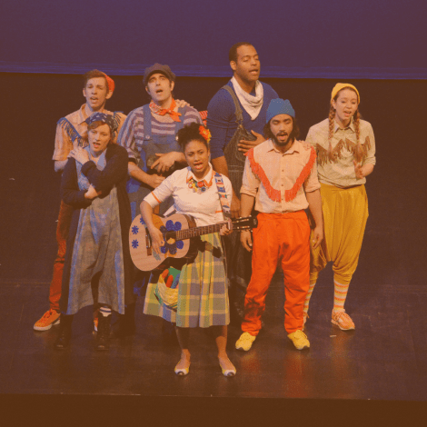 A group of performers in eclectic clothing, sing on a stage. A woman in front holds a guitar.